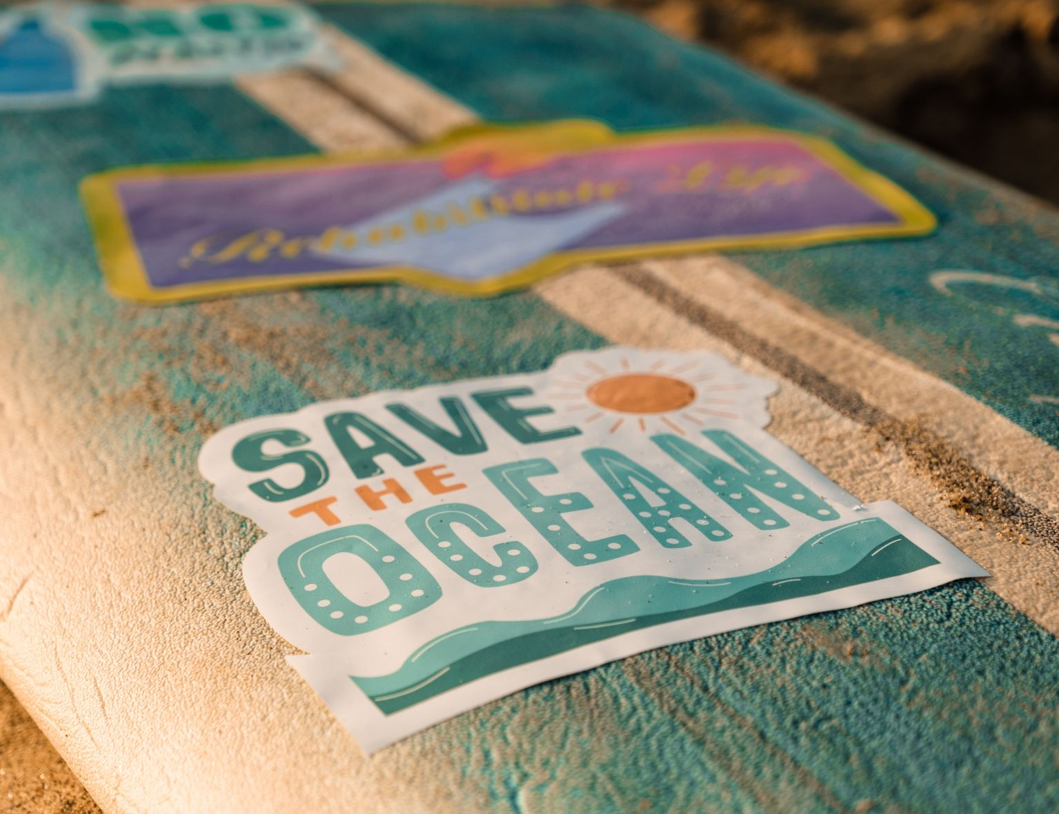 Ein Surfbrett mit einem "SAVE THE OCEAN"-Aufkleber mit Sonne und Wellen auf einem Sandstrand, betont das Thema Ozeanschutz.