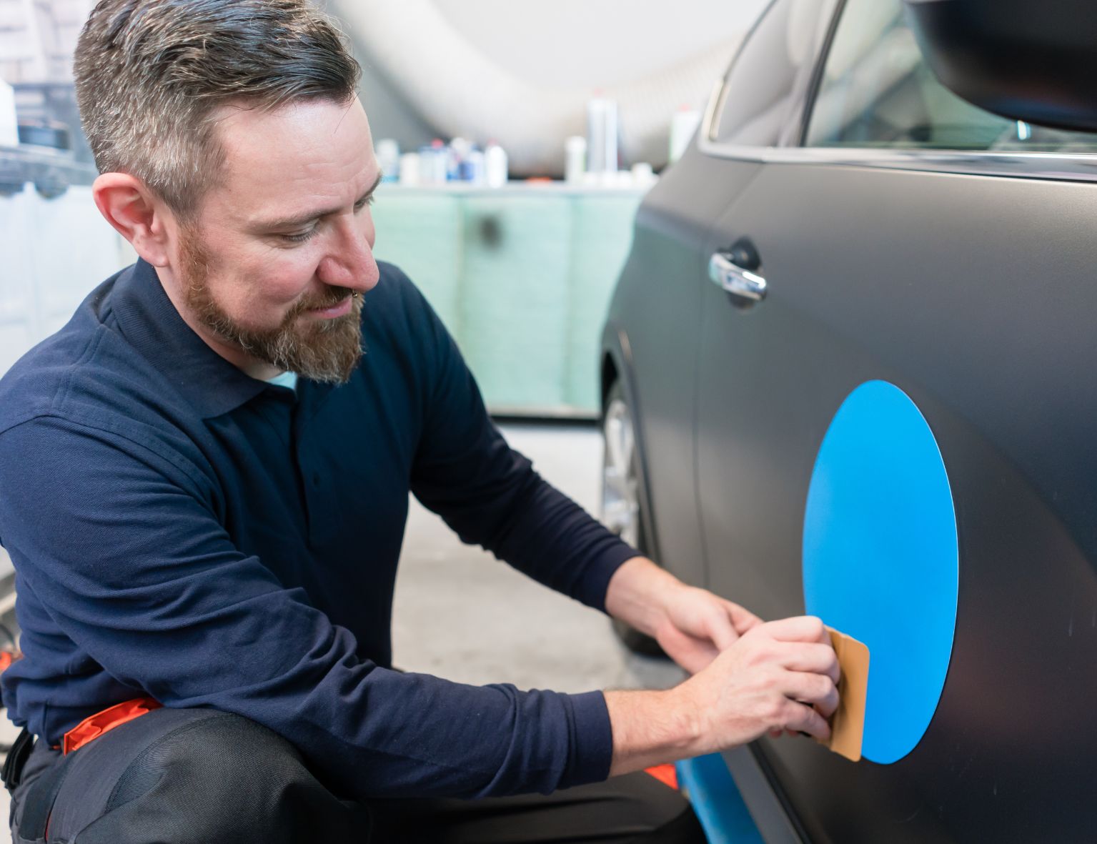 Mann, der eine blaue Vinyl-Autofolie auf eine schwarze Autotür aufträgt und sie mit einem Werkzeug glättet, umgeben von einer Werkstattumgebung.