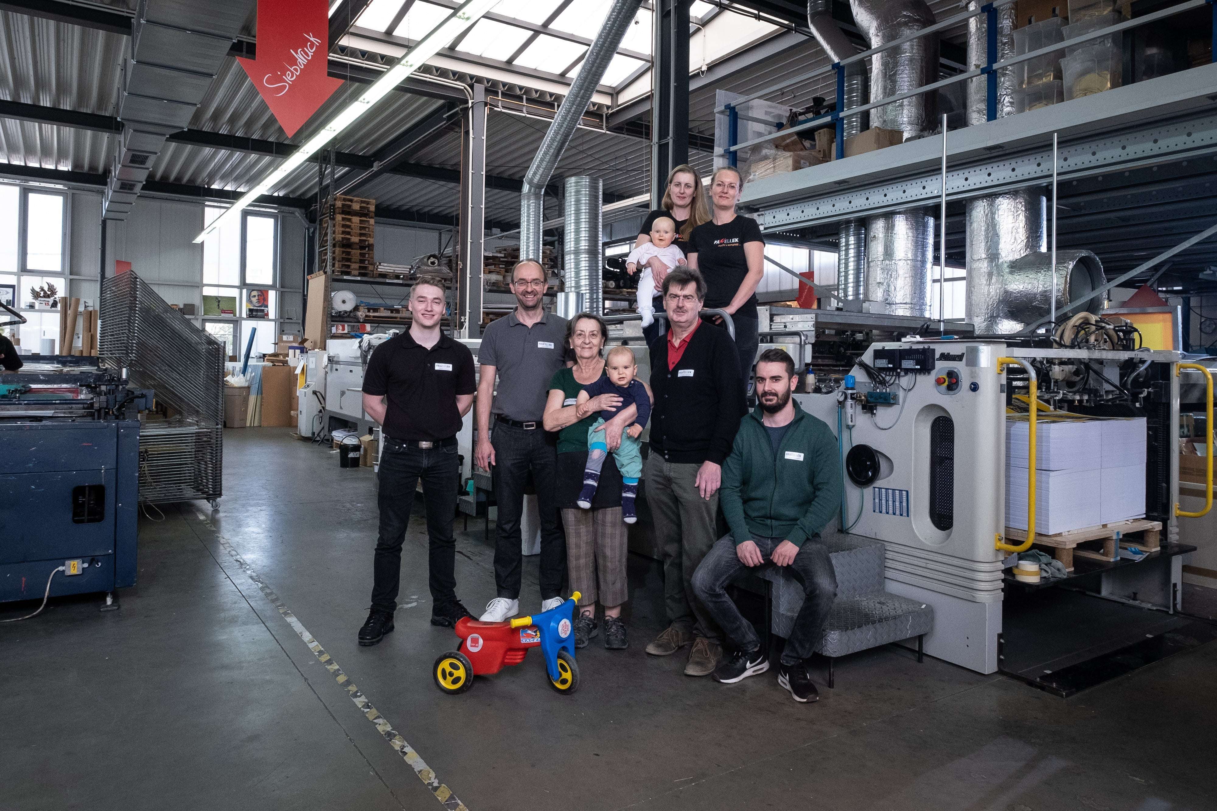 Gruppe von Menschen in einer industriellen Umgebung mit Maschinen, einem roten Schild und einem bunten Spielzeug-Lkw.