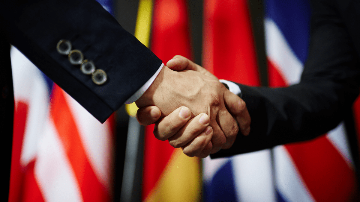 Zwei Personen schütteln sich die Hände vor mehreren internationalen Flaggen, was globale Zusammenarbeit symbolisiert.