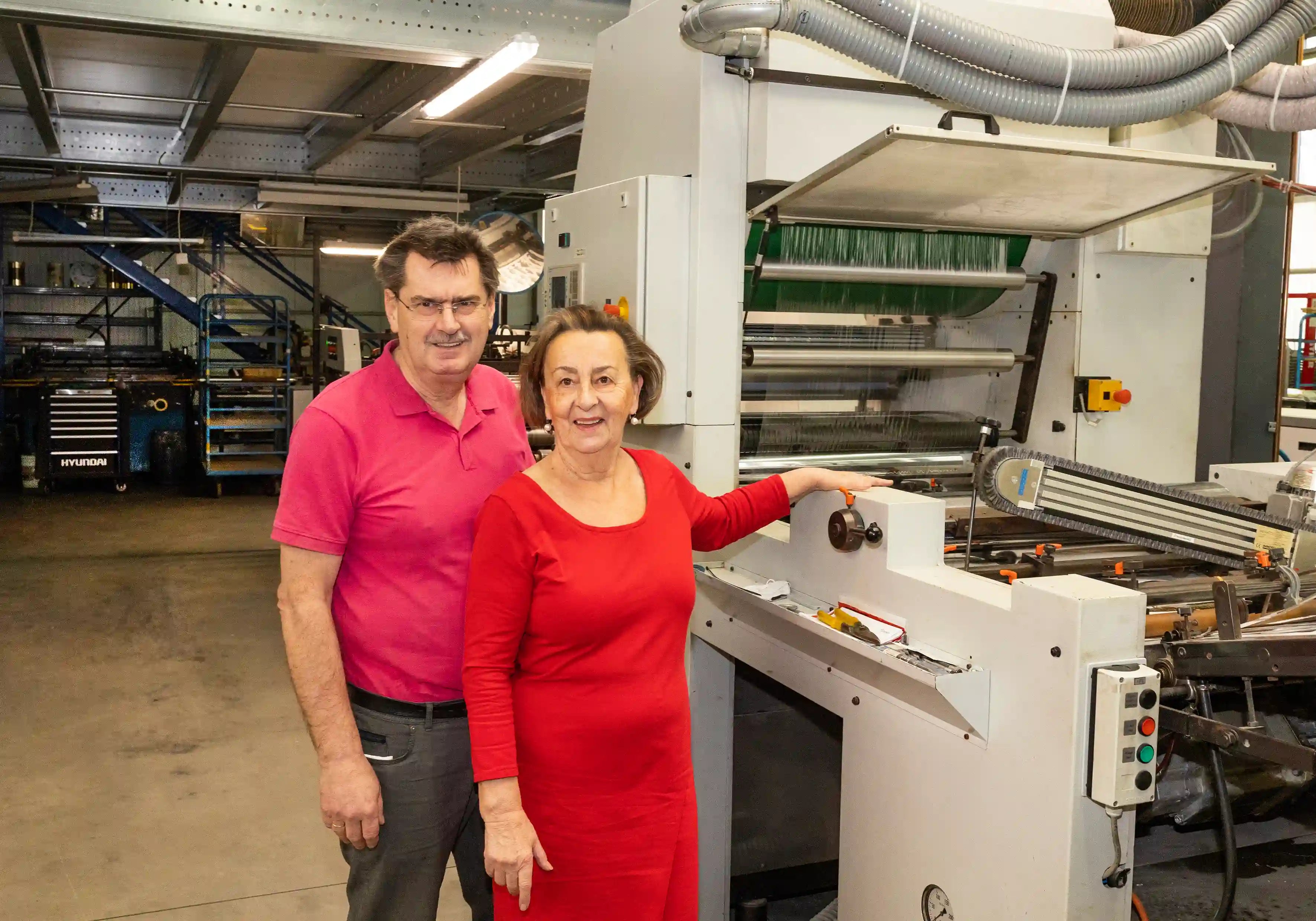 Zwei Personen, die neben einer großen Industriemaschine in einer Werkstatt oder einer Fertigungsumgebung stehen.