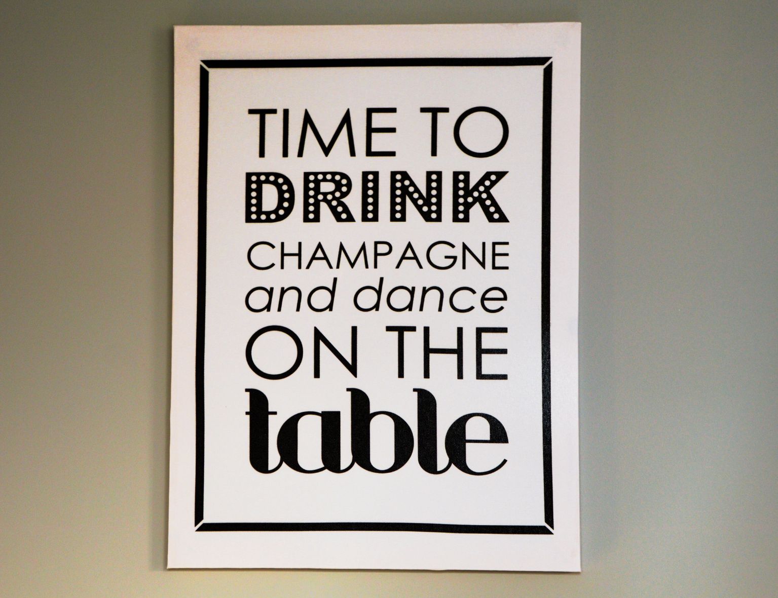 Ein gerahmtes Bild mit dem Text: "Time to drink champagne and dance on the table." Schlichtes, modernes Design auf hellem Hintergrund.