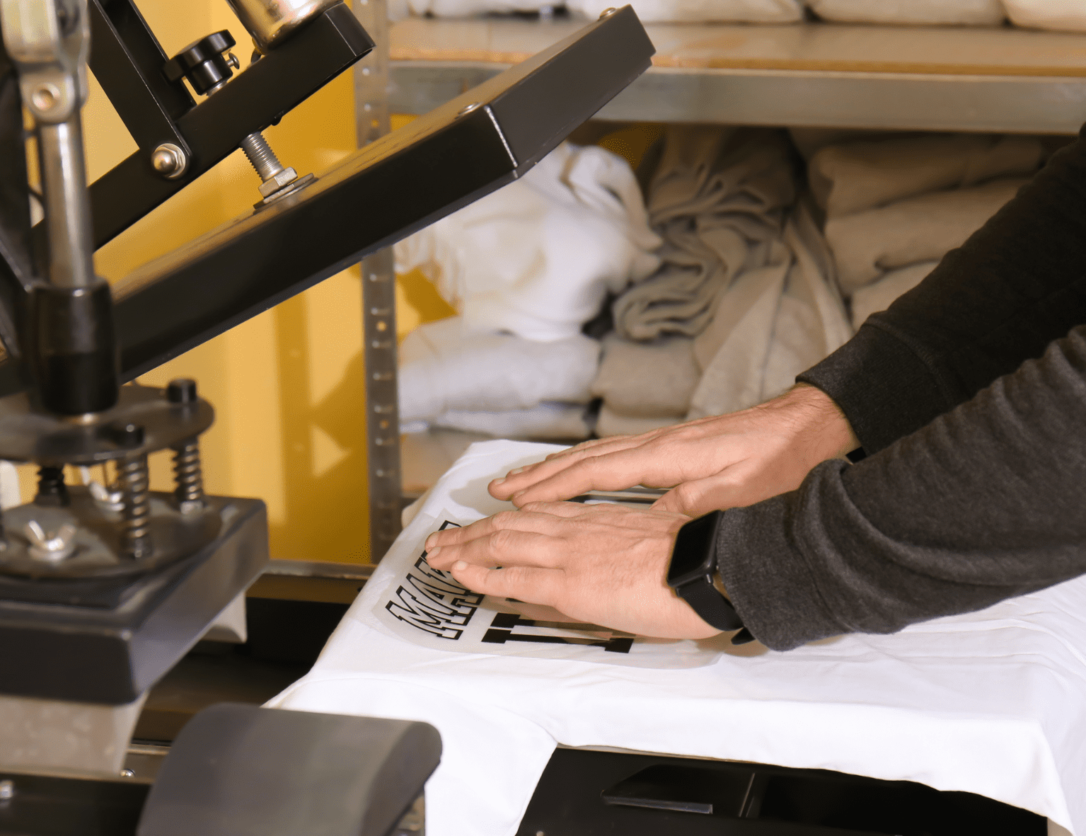 Hände pressen Design auf weißes T-Shirt mit Transferpresse in Textilstudio, umgeben von Stoffen, betont T-Shirt-Druck.
