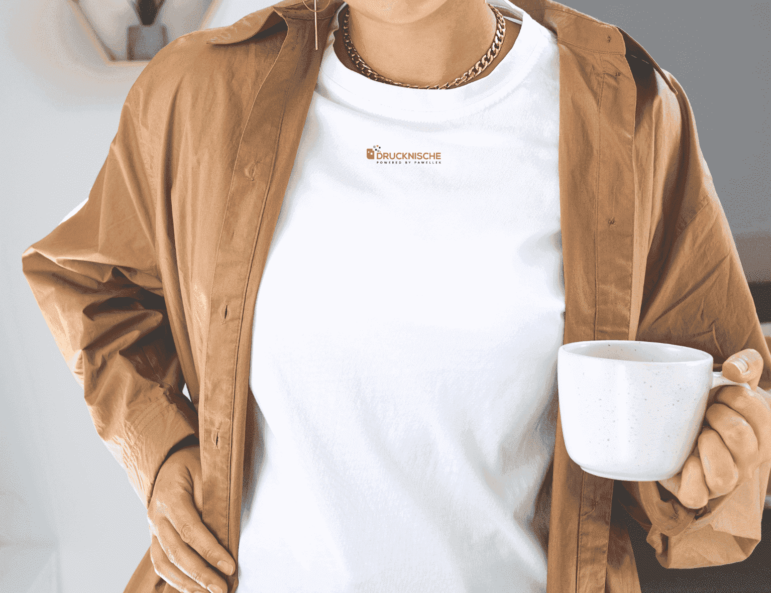 Eine Person trägt ein weißes T-Shirt mit kleinem Logo unter einer braunen Jacke und hält eine weiße Tasse in der Hand.