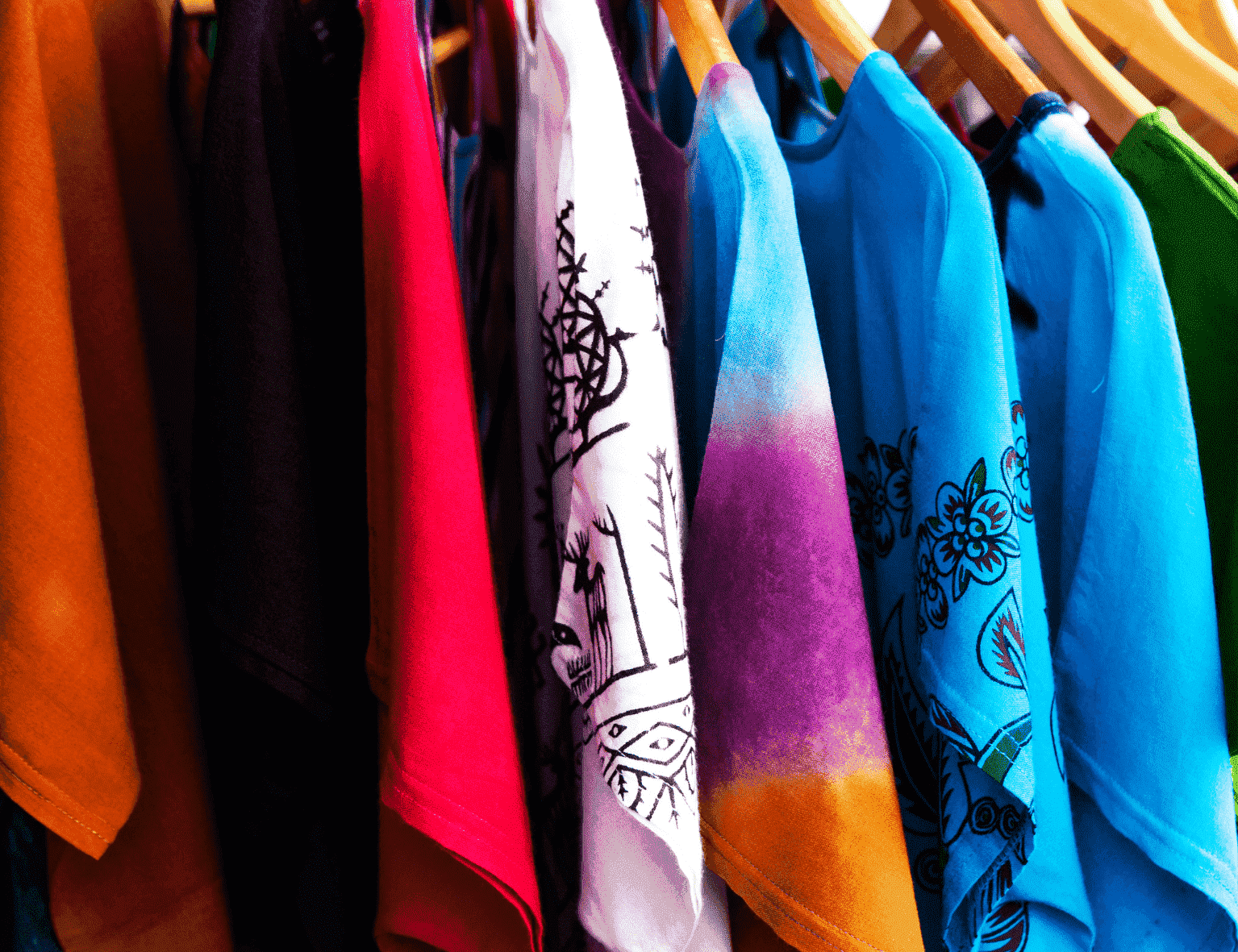 Auf einem Ständer hängen farbenfrohe, gemusterte T-Shirts mit Motiven wie Blumendrucken und Tie-Dye-Effekten.