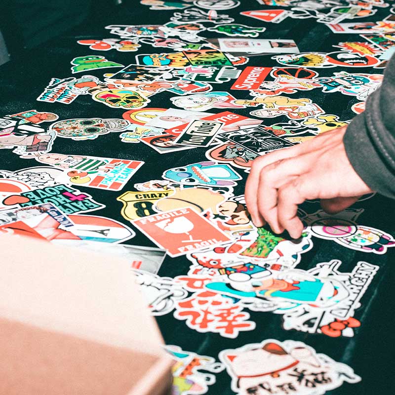 Ein Tisch voller bunter Aufkleber mit verschiedenen Designs; eine Hand wählt einen aus; kreative Auswahl an Stickern.
