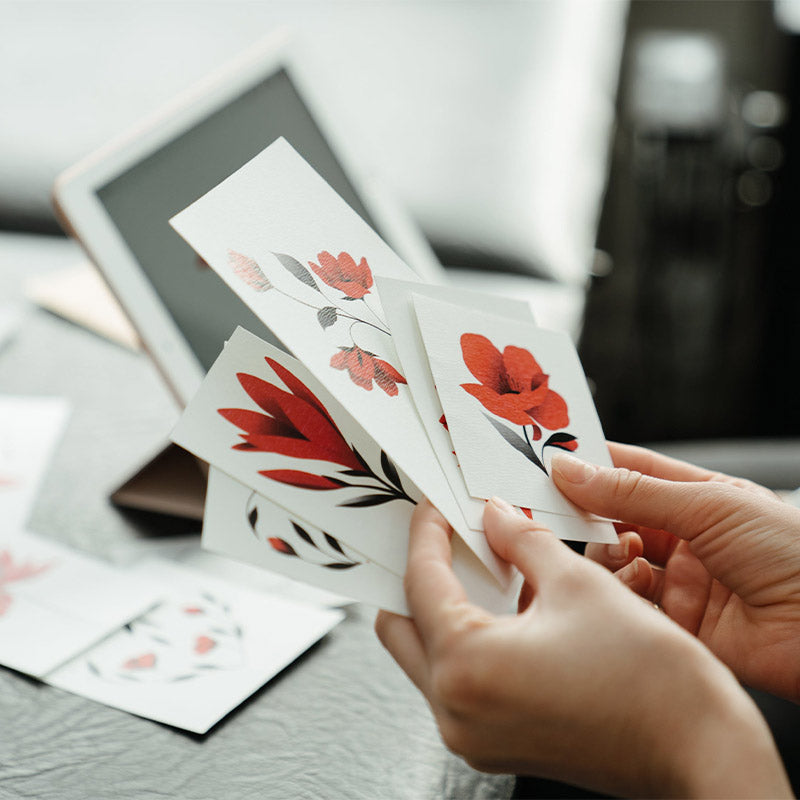 Person hält Karten mit Blumenmuster in Rot und Schwarz, sitzt neben einem Tablet auf grauer, strukturierter Oberfläche.