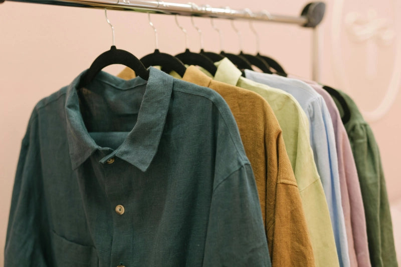 Eine Reihe von Leinenhemden auf Kleiderbügeln, gezeigt in verschiedenen Farben wie Blau, Braun, Grün, Lila und Rosa.