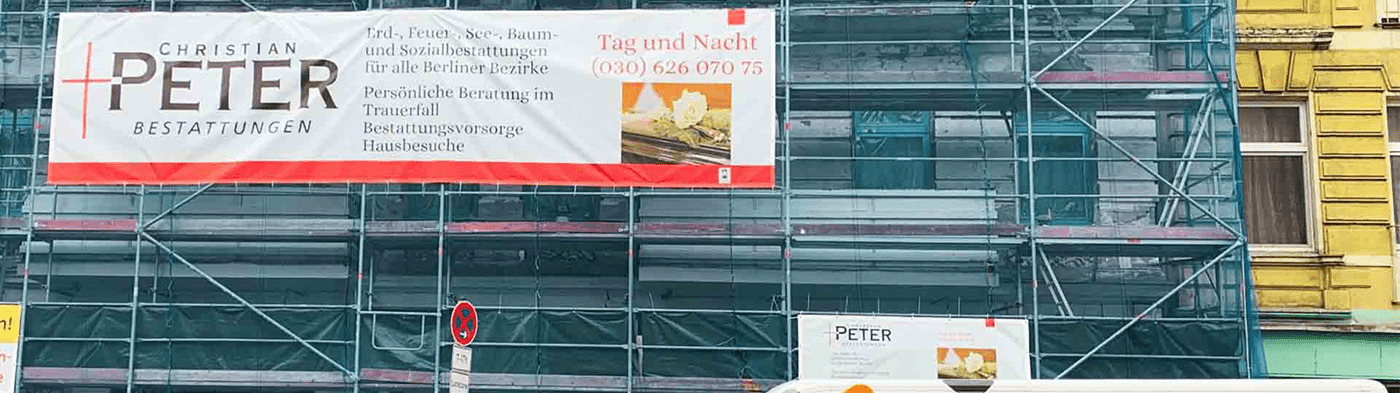 Ein Baugerüst vor einem Gebäude mit einem großen Schild für "Christian Peter Bestattungen", das Bestattungsdienste bewirbt. Unten steht ein Fahrzeug mit ähnlicher Beschriftung.