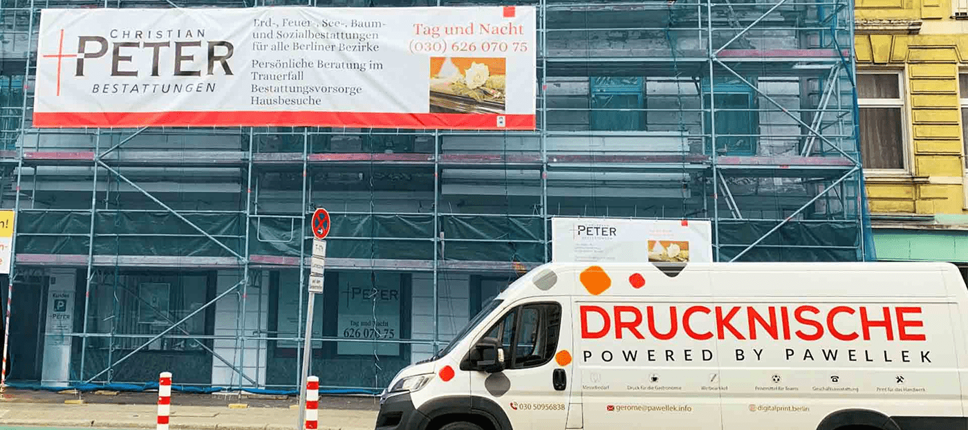 Ein weißer Van mit "DRUCKNISCHE"-Branding steht vor einem Gebäude mit Baugerüst, das ein Bestattungsunternehmen-Banner zeigt.