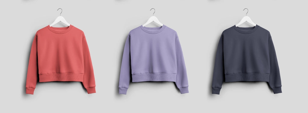 Drei modische Sweatshirts mit Rundhalsausschnitt in Koralle, Lavendel und Dunkelgrau, die auf Kleiderbügeln vor einem neutralen Hintergrund präsentiert werden.