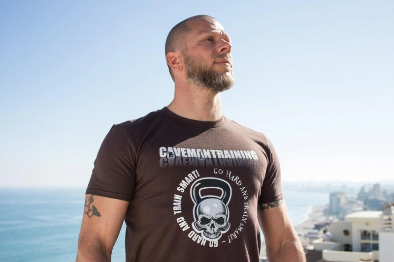 Ein Mann mit Bart trägt ein braunes T-Shirt mit dem Aufdruck "Cavemantraining" und einem Totenkopf. Er steht im Freien vor einem klaren Himmel und Blick aufs Meer.