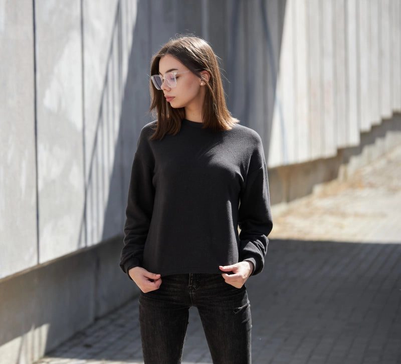 Eine Frau trägt einen schwarzen Pullover und Jeans, steht in urbaner Umgebung mit Betonwänden, Sonnenbrille tragend, bei Sonnenlicht.