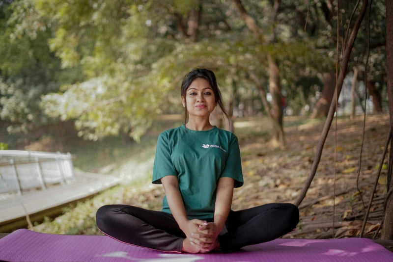Eine Person sitzt mit gekreuzten Beinen auf einer lila Yogamatte im Freien, umgeben von Bäumen und Grün. Sie lächelt und trägt ein grünes T-Shirt und schwarze Leggings.