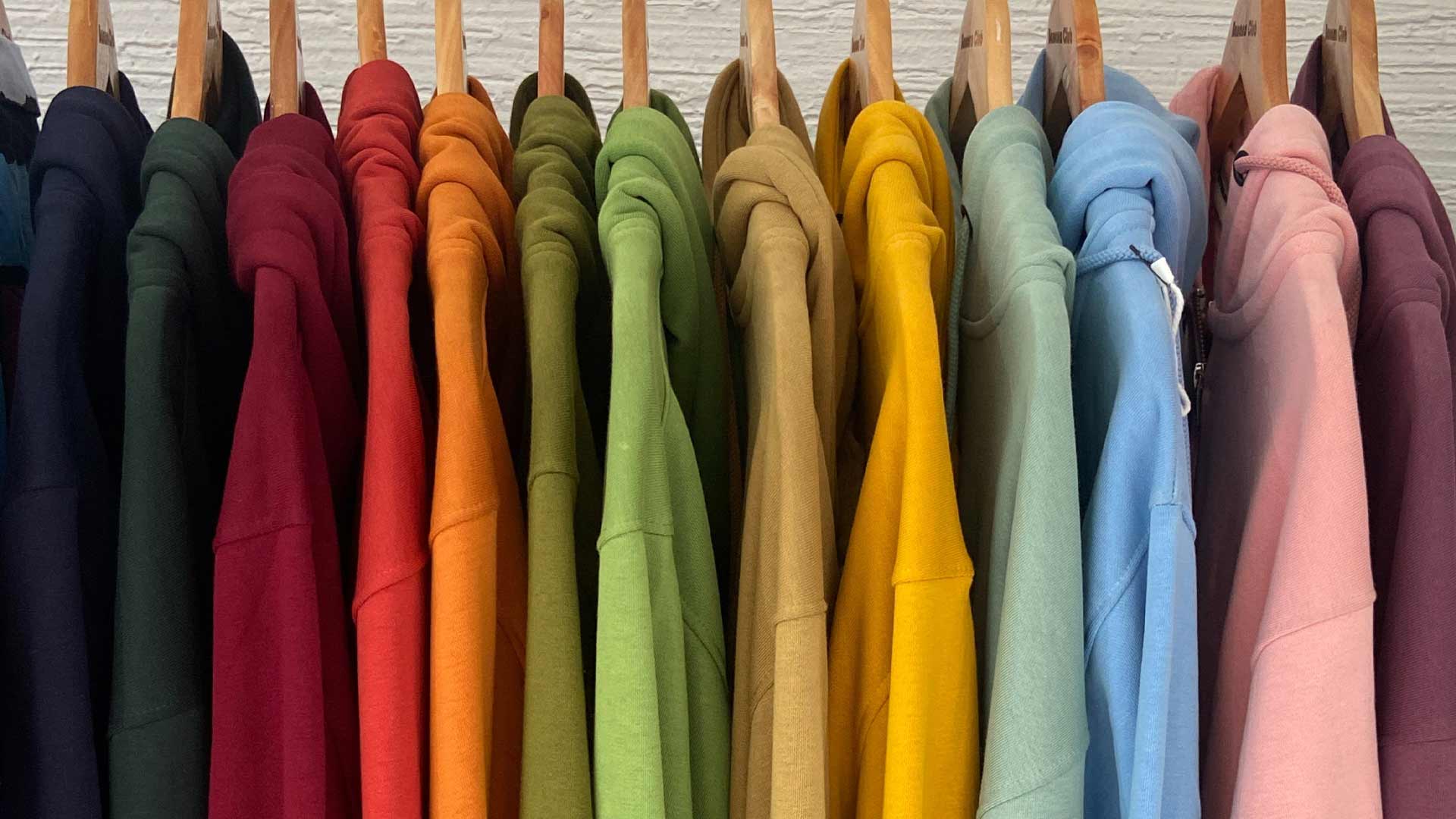 Eine Reihe von bunten Kapuzenpullovern in verschiedenen Farben, die ordentlich an Holzkleiderbügeln aufgehängt sind.