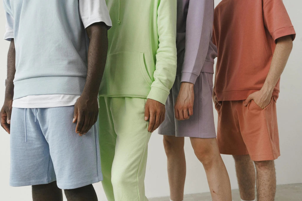 Vier Personen in pastellfarbenen Shorts und Sweatshirts; Blau-, Grün-, Lavendel- und Korallentöne; lässiger Modestil.