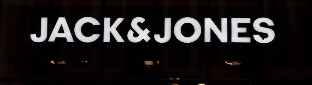 Ein hell erleuchtetes weißes „JACK & JONES“-Geschäftsschild auf dunklem Hintergrund, das das Markenlogo in einer fetten, modernen Schrift hervorhebt.