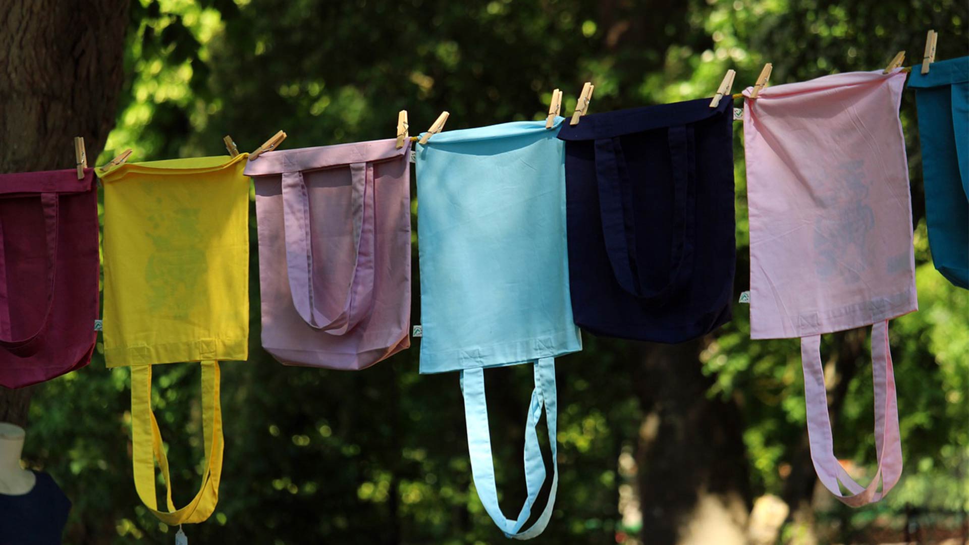 Bunte Stofftaschen hängen an einer Wäscheleine im Freien, festgehalten mit Holzklammern; grüner, verschwommener Hintergrund.