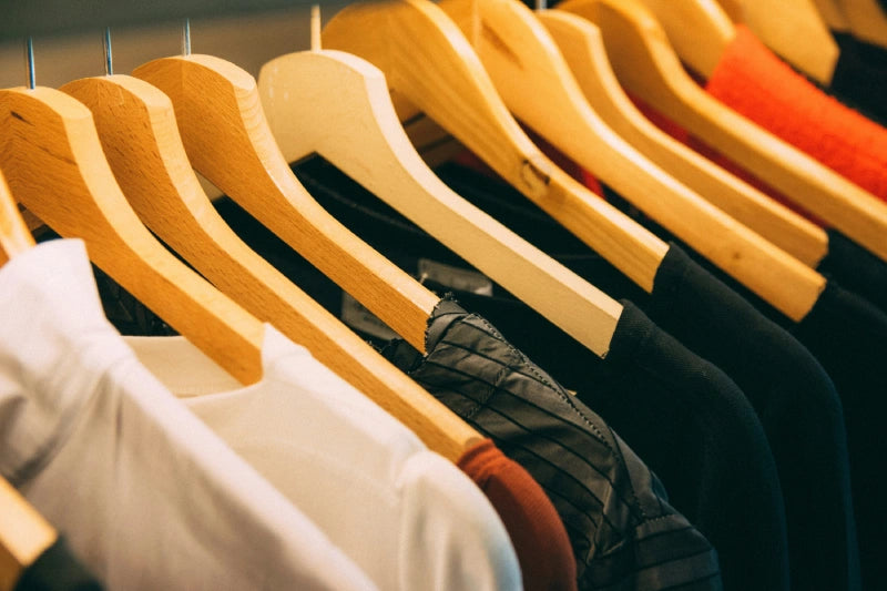 Ein Kleiderbügel auf einer Stange mit ordentlich gehängten Kleidung, darunter weiße Hemden, schwarze Jacken und bunte Teile.