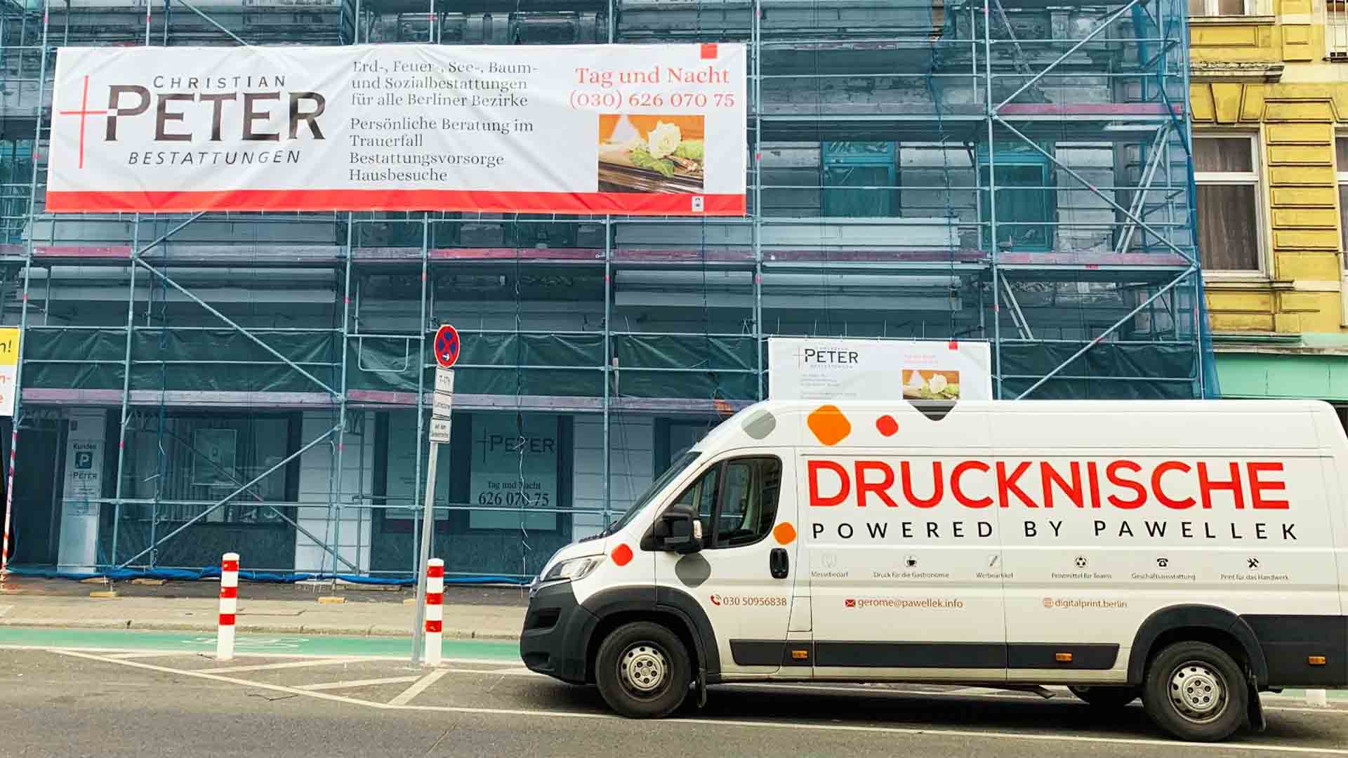 Eine Baustelle mit eingegerüstetem Gebäude, Banner mit "Bestattungen", weißer Lieferwagen mit "DRUCKNISCHE" Beschriftung.