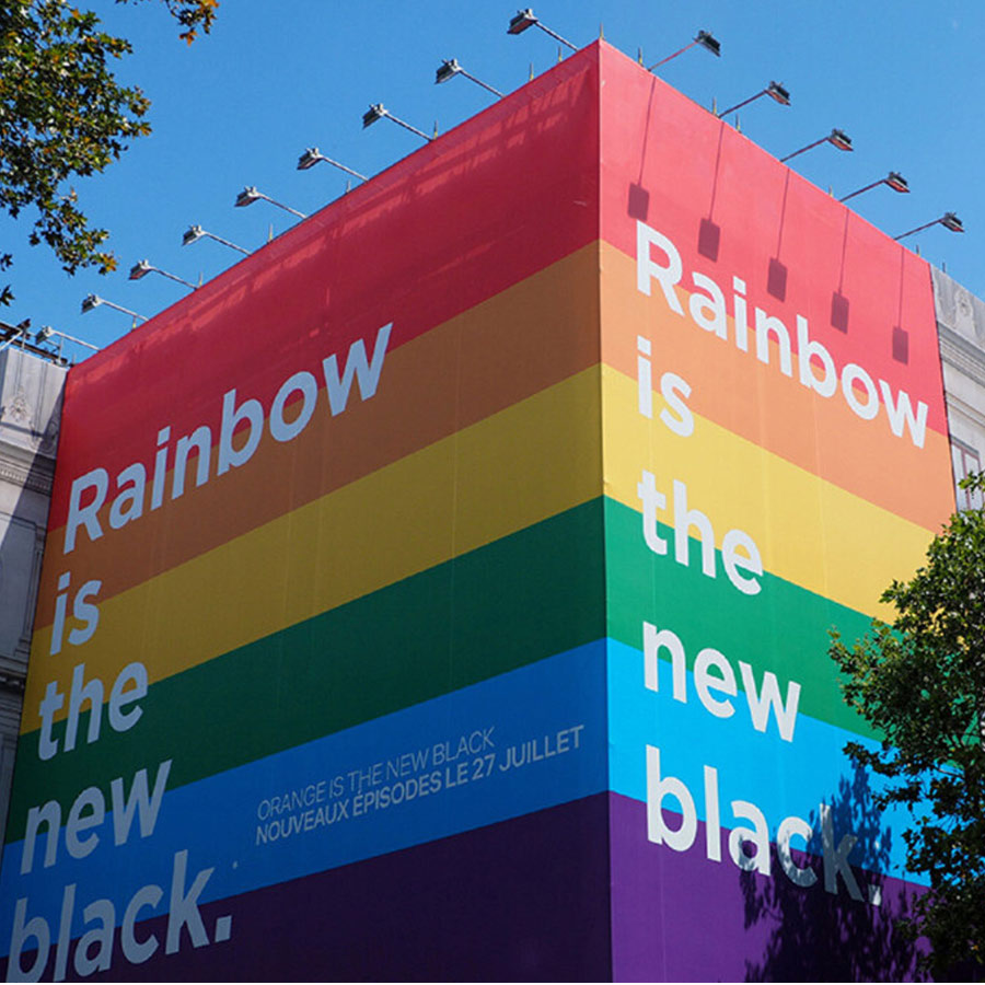 Ein großes Wandgebäude mit Regenbogenstreifen und dem weißen Text "Rainbow is the new black" unter blauem Himmel.