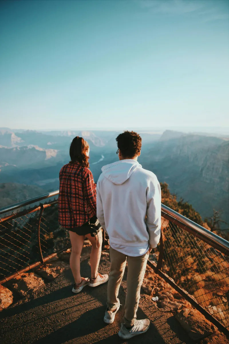 Zwei Personen stehen an einem Aussichtspunkt und blicken auf eine weite, bergige Landschaft unter klarem blauem Himmel.