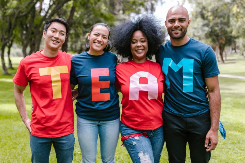 Personen stehen in einem Park und tragen bunte T-Shirts, die das Wort "TEAM" buchstabieren.