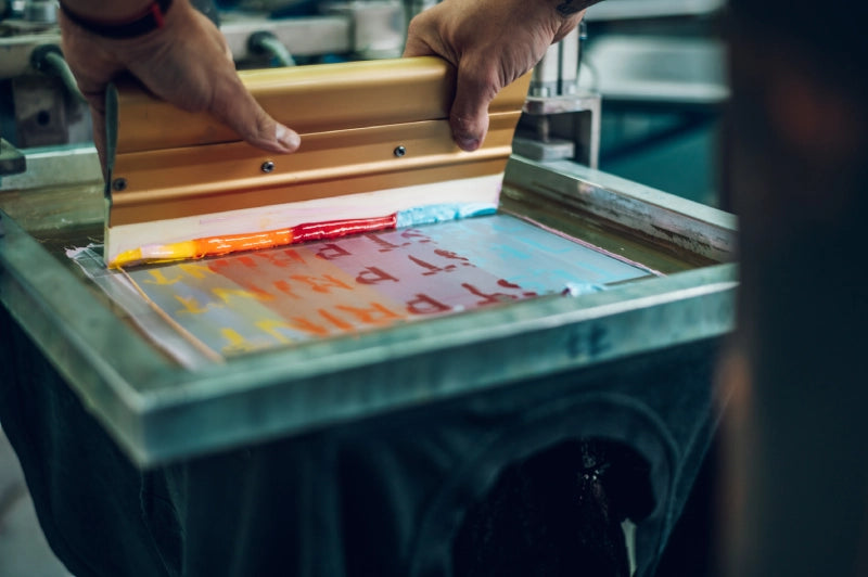 Eine Person verwendet einen Rakel, um bunte Tinte auf einen Siebdruckrahmen für das Bedrucken von T-Shirts aufzutragen.
