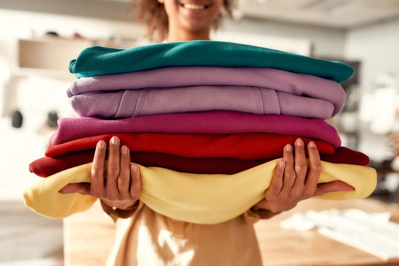 Eine Person präsentiert einen Stapel sorgfältig gefalteter Pullover in lebhaften Farben, darunter Rot, Lila, Gelb und Grün.