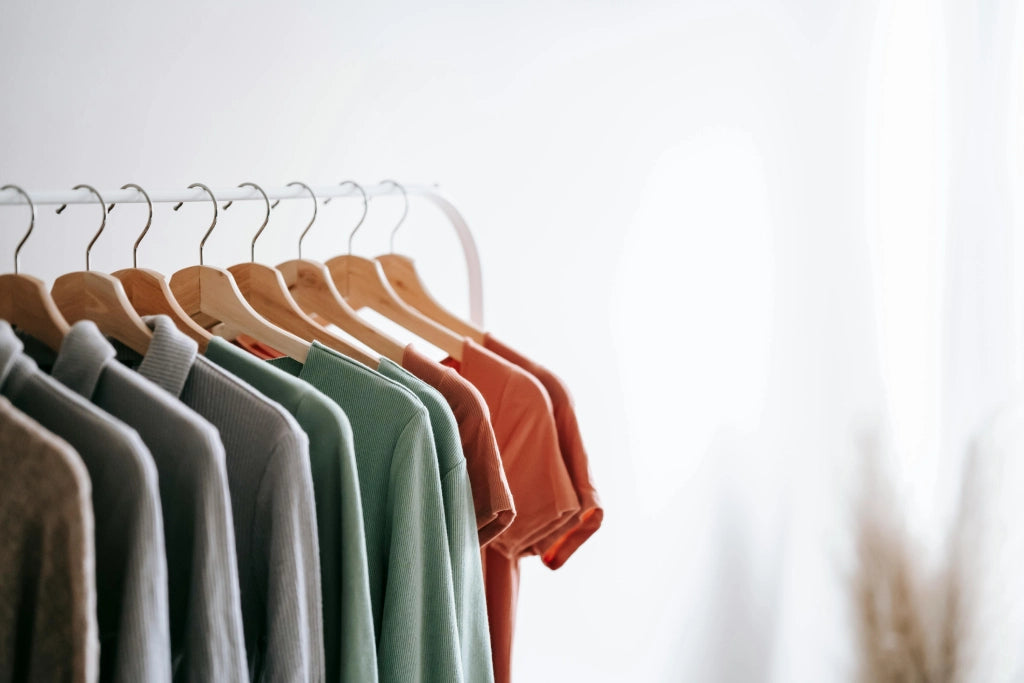 Ein Kleiderständer mit verschiedenen pastell- und erdfarbenen T-Shirts auf Holzbügeln vor einem weichen, minimalistischen Hintergrund.