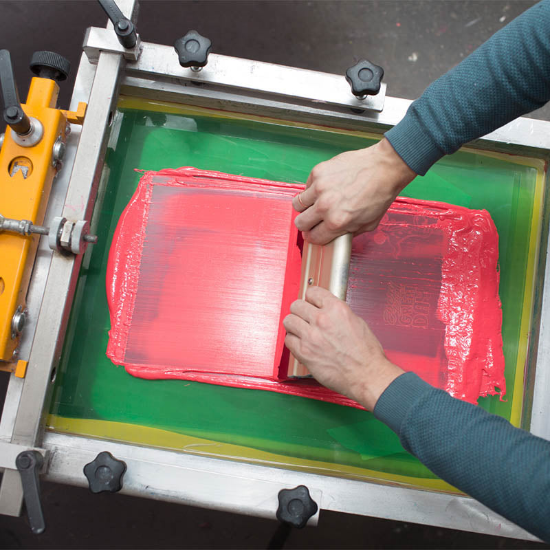 Siebdruckprozess, bei dem rote Tinte mit einem Holzrakel auf einem grünen Sieb gleichmäßig verteilt wird.