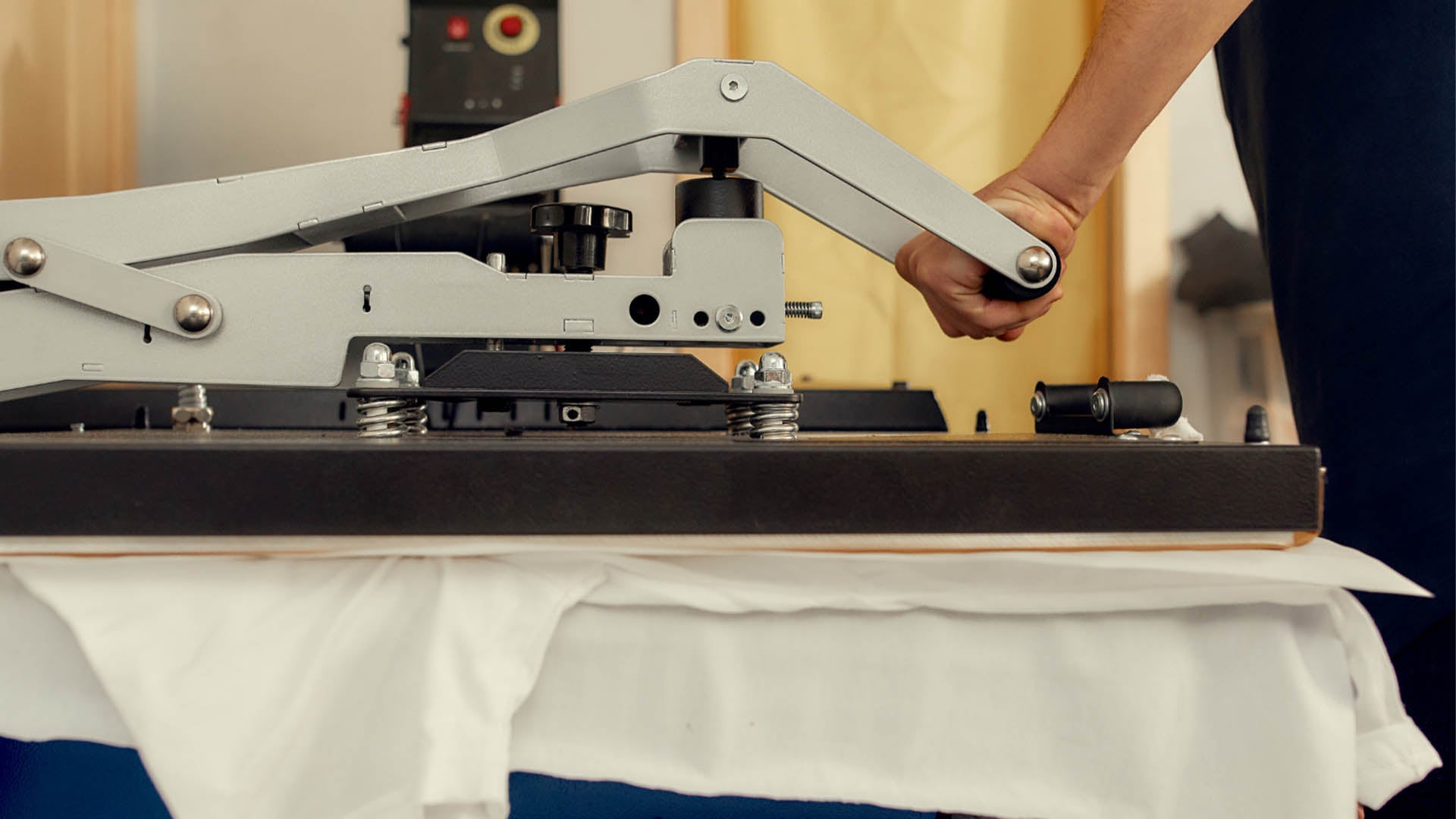 Eine Wärmepresse, die mit einem von einer Person gehaltenen Hebel bedient wird, der über einem sauber platzierten weißen Stoff liegt.