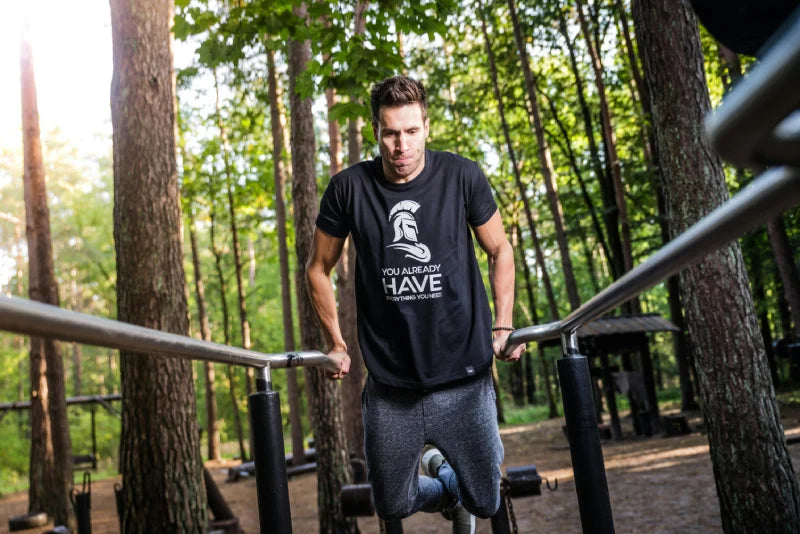 Ein Mann trainiert an Parallelstangen im Wald, trägt ein schwarzes T-Shirt mit Motiv und graue Hose, während Sonnenlicht durch die Bäume scheint.