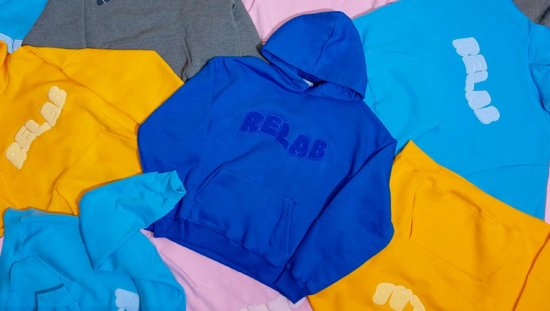 Verschiedene farbige Hoodies in Blau, Gelb, Grau und Pink mit dem Aufdruck "RELAP" flach auf einer Oberfläche ausgelegt.