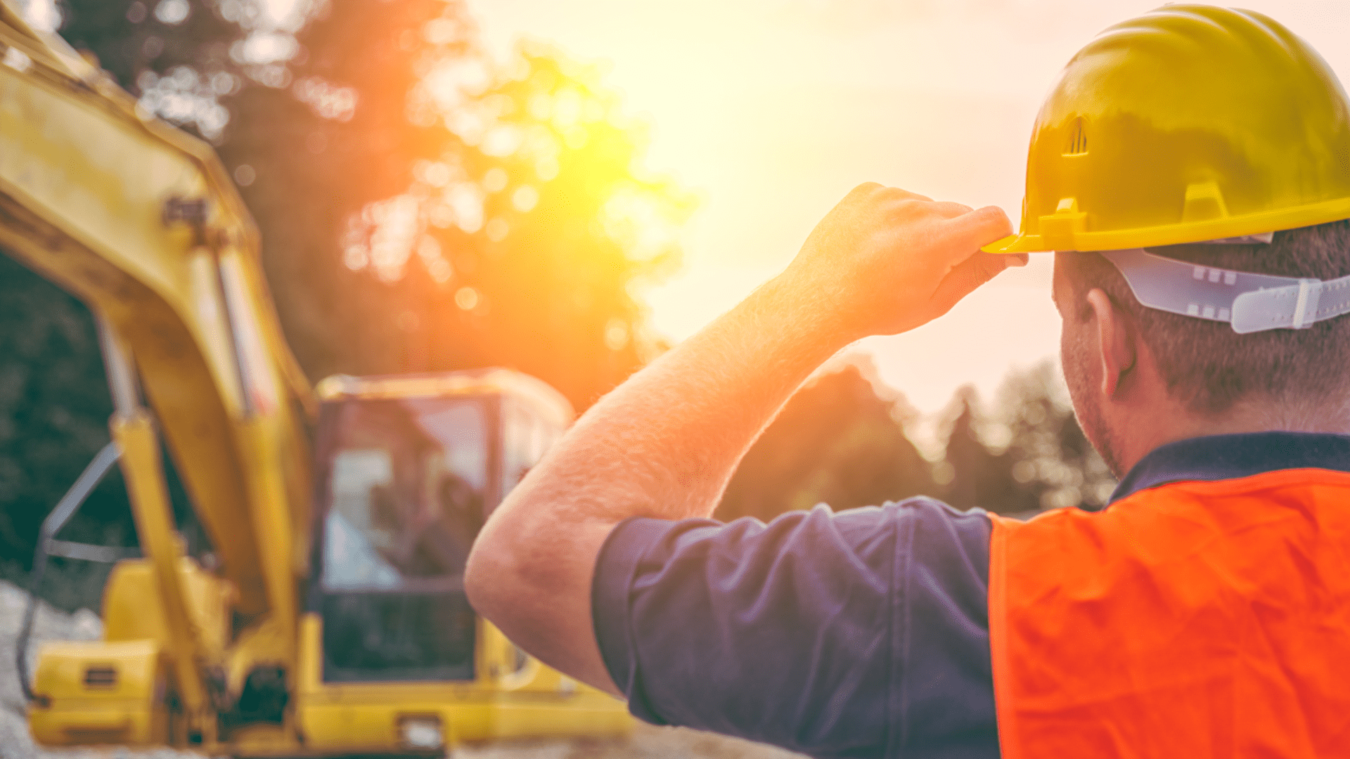 Ein Bauarbeiter mit gelbem Schutzhelm und orangefarbener Weste, der auf eine Baustelle mit Bagger bei Sonnenuntergang blickt.